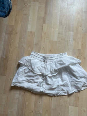 Vit volangkjol med knytband - Kort vit kjol i luftig linnemix med bred resår i midjan och dekorativt knytband med tofsar. Kjolen har volangdetalj upptill som ger en söt och avslappnad look. Perfekt för varma dagar och enkel att matcha med olika toppar.