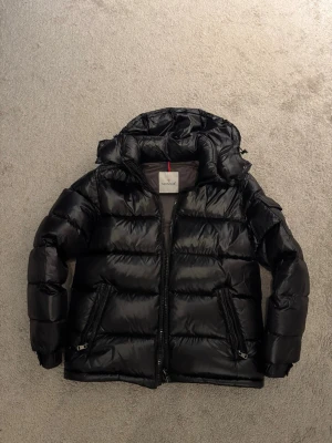 Svart pufferjacka från Moncler - Snygg svart pufferjacka från Moncler med glansig finish och huva. Jackan har två dragkedjeförsedda fickor framtill och en hög krage. Perfekt för kalla vinterdagar och riktigt trendig look.
