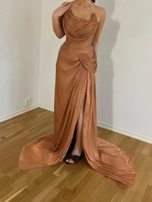 Lång bronsfärgad klänning med slits - Miss Rosier maxiklänning – Ny med prislapp ✨  Säljer en helt ny och oanvänd maxiklänning från Miss Rosier, fortfarande med prislappen kvar.  Den har en elegant draperad design med en smickrande passform, hög slits och ett vackert släp – perfekt till bal, gala, bröllop eller andra festliga tillfällen.  Köpte för 2500 kr men kan släppa för 1500kr. Originalpris: 2 500 kr 💰 Säljes för: 1 500 kr  📏 Storlek: S (36) 🏷️ Skick: Helt ny med prislapp 🎨 Färg: Guld/Brun