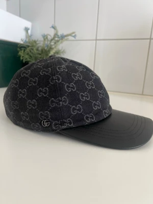 Svart keps med GG-mönster från Gucci - Snygg svart keps från Gucci med klassiskt GG-monogram i grått över hela tyget. Skärmen är i svart läder och kepsen har en metallogo på sidan. Perfekt accessoar för dig som gillar exklusiv streetwear och vill sticka ut med en ikonisk detalj.