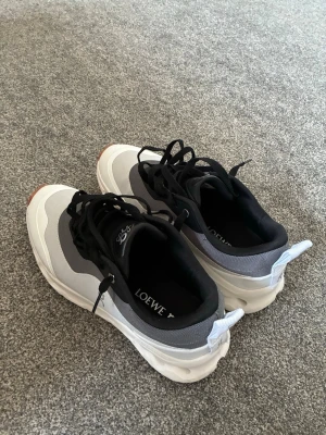 Loewe x On Running sneakers grå/vit - Snygga sneakers från Loewe x On Running i grått och vitt med svarta detaljer. Skorna har en chunky sula med hål och svarta skosnören. Ovandelen är i mesh och syntetmaterial för en sportig look. Perfekta för dig som gillar streetwear och unika samarbeten.