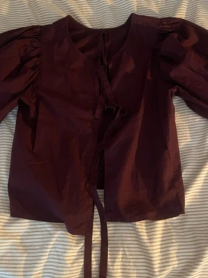 Vinröd blus med puffärmar och knytband - Säljer en vinröd blus med puffiga långärmar och knytband framtill. Blusen har rund halsringning och är gjord i ett vävt tyg som känns lite styvt. Perfekt för dig som vill ha en statement-blus med coola detaljer.