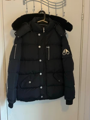 Svarta Mosse knuckles - Snygg svart dunjacka med stor huva och pälskant. Jackan har silverfärgade knappar och dragkedjor, samt en logga på ärmen. Flera praktiska fickor och quiltad design gör den både varm och stilren. Perfekt för kalla vinterdagar.