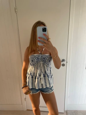 Vit och blå bandeau topp med mönster - Säljer en söt bandeau topp i vitt med blått broderat mönster. Toppen har en bandeauform med knyt i mitten och tunna axelband. Perfekt för sommaren!