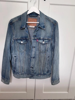 Levi's klassisk blå jeansjacka - Säljer en klassisk blå jeansjacka från Levi's med två bröstfickor med lock och knappar, samt sidofickor. Jackan har en tidlös tvättad look, raka sömmar och silverfärgade knappar. 
