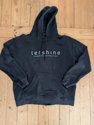 Svart hoodie från Tershine - Svart hoodie från Tershine med vit text 'swedish perfection' på bröstet. Klassisk känguruficka framtill och justerbar huva med snören. Tillverkad i mjukt material, perfekt för en avslappnad och stilren look.