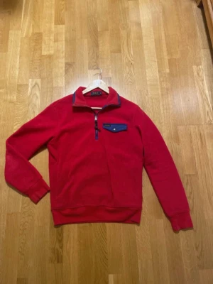 Polo Ralph Lauren röd fleece-tröja Strolek S - Röd långärmad fleece-tröja från Polo Ralph Lauren med halv dragkedja och hög krage. Bröstficka med blå kant och knapp, samt blå detaljer vid dragkedjan och kragen. Ribbade muddar vid ärmslut och nederkant. Perfekt för lager-på-lager under hösten och vintern. Väldigt bra skick, knappt använd. Storlek Small
