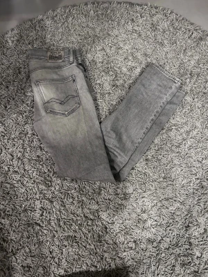 Grå slim fit jeans från Replay - Snygga grå slim fit jeans från Replay med klassisk femficksdesign och diskreta slitningar. Jeansen har en ljusgrå tvätt och tydliga sömmar på bakfickan. Perfekta för en trendig och avslappnad look.