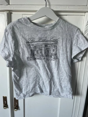 Grå t-shirt med london motiv - Säljer en ljusgrå t-shirt med korta ärmar och ett tryck framtill som föreställer en butik med texten 'The West End'. T-shirten har rund halsringning och är i mjukt bomullsmaterial. Perfekt för en avslappnad och trendig look.
