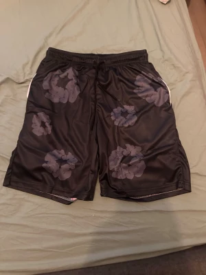 Svarta shorts med blommigt mönster - Säljer ett par svarta shorts med stora blommiga mönster i mörkgrått. Shortsen har resår i midjan med snörning och är gjorda i ett glansigt syntetmaterial. Perfekta för dig som gillar en avslappnad och trendig stil.