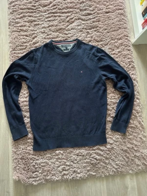 Mörkblå stickad tröja Tommy Hilfiger - Snygg mörkblå stickad tröja från Tommy Hilfiger i premiumkvalitet med pima cotton och cashmere. Tröjan har rund halsringning, diskret logga på bröstet och ribbade muddar vid ärmslut och nederkant. Perfekt för en stilren look.