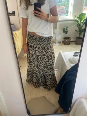 Långkjol med mönster från Zara - Säljer en flowy svartvit långkjol med elastisk midja och grafiskt mönster. Kjolen har volang nedtill och är perfekt för dig som gillar en bohemisk och avslappnad stil. Passar till både sneakers och sandaler.