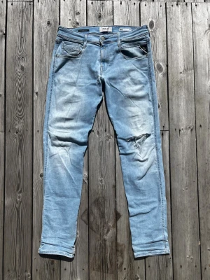 Replay Anbass jeans ljusblå - Ett par jeans i ljusblå fadead tvätt från Replay. Schysst tvätt på framsidan samt en cool slitning på vänster knä. Jeansen är i ett riktigt najs stretch material. Storleken är W30 och L32. Modellen är slim fit.
