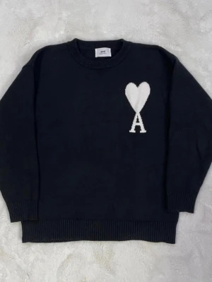 Ami Paris stickad tröja - Mörkblå stickad tröja från Ami med rund halsringning och ribbade muddar. På bröstet finns en vit hjärtdetalj med bokstaven 'A' under, vilket ger en unik och trendig look. Perfekt för dig som gillar stilrena plagg med en twist.