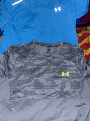Två Under Armour tränings-tshirts - Säljer två snygga tränings-tshirts från Under Armour. Ena är blå med vit logga och den andra är grå med camomönster och gul logga. Perfekta för gymmet eller löprundan, båda har rund hals och är tillverkade i funktionsmaterial som andas.