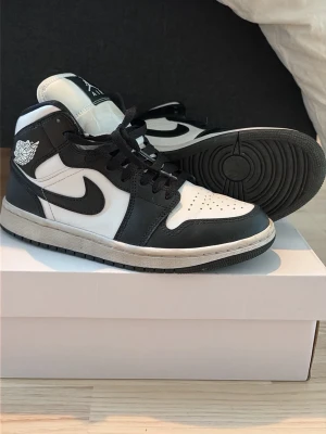 Svarta och vita Air Jordan 1 Mid - Säljer ett par klassiska Air Jordan 1 Mid sneakers i svart och vitt läder. Skorna har svart swoosh, svarta snören och vita detaljer på tån och plösen. Ikonisk Jordan-logga på sidan och Jumpman på plösen. 