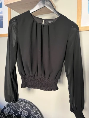 Svart blus med smockad midja - Stilren svart blus med långärmad design och rund halsringning. Blusen har smockad midja och är tillverkad i ett tunt, lätt material som ger ett snyggt fall. Perfekt att matcha med både jeans och kjol för en trendig look.