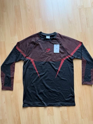 Onyx V5 long sleeve  - Säljer en svart långärmad träningströja från Gymshark med röda detaljer och mönstrade partier på axlar och ärmar. Tillverkad i ett lätt och stretchigt syntetmaterial som andas, perfekt för gym eller löpning.