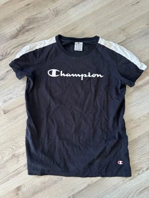 Svart Champion t-shirt med vita detaljer - Svart t-shirt från Champion med vita partier på axlarna och stort Champion-tryck över bröstet. Klassisk rund hals och korta ärmar. Perfekt för en sportig och avslappnad stil.