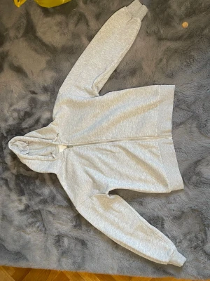 Ljusgrå hoodie från Gina Tricot - Ljusgrå hoodie från Gina Tricot med dragkedja framtill och justerbar huva med snören. Tillverkad i mjukt sweatshirtmaterial och har ribbade muddar vid ärmar och nederkant. Perfekt för en chill och avslappnad stil.