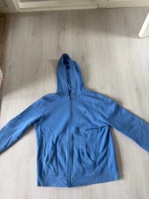 Blå hoodie från Polo Ralph Lauren - Snygg blå hoodie från Polo Ralph Lauren med dragkedja framtill och två fickor. Klassisk huva och diskret logga broderad på bröstet. Tillverkad i mjukt bomullsmaterial som är skönt att ha på sig. Perfekt för en chill och avslappnad stil.