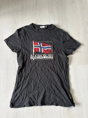 Svart t-shirt från Napapijri - Svart t-shirt från Napapijri med tryck av norska flaggan och logga på bröstet. Klassisk rund hals och korta ärmar. Tillverkad i mjuk bomull som känns skön mot huden. Perfekt för en avslappnad och stilren look.