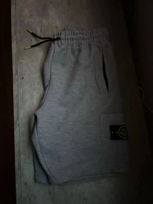 Grå shorts med snörning Stone Island - Säljer ett par grå shorts från Stone Island med elastisk midja och svart snörning med gulddetaljer. Shortsen har två sidofickor och en ficka med Stone Island-logga på benet. Perfekta för chill dagar eller streetwear-stil.