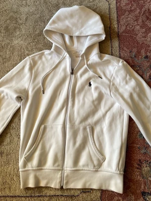 Vit hoodie från Polo Ralph Lauren - Snygg vit hoodie från Polo Ralph Lauren med dragkedja framtill och två fickor. Klassisk logga broderad på bröstet och justerbar huva med snören. Tillverkad i mjuk bomull. 8/10 skick. Skriv gärna om frågor!