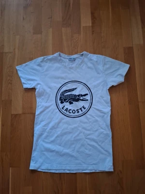 Vit t-shirt från Lacoste med logga - Säljer en vit t-shirt från Lacoste med stor svart logga och krokodiltryck på bröstet. Klassisk rund hals och korta ärmar. Perfekt till jeans eller shorts för en clean och sportig look.