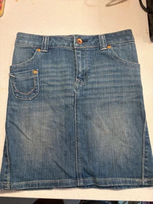 Blå jeanskjol med fickor - Säljer en klassisk blå jeanskjol med fem fickor och bälteshällor. Kjolen har en knapp och dragkedja framtill samt dekorativa sömmar och en snygg patch bak. Perfekt till vardags för en avslappnad look.