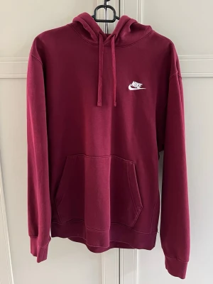 Vinröd hoodie från Nike med logga - Snygg vinröd hoodie från Nike med klassisk vit logga på bröstet. Hoodien har huva med dragsko, stor magficka och ribbade muddar vid ärmar och nederkant. Perfekt för en chill och sportig look.