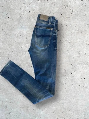 Blå slim fit jeans med slitningar -  Feta nudie.Snygga blå slim fit jeans med slitningar och tvättade detaljer. Klassisk femficksmodell med gylf och knapp, samt en mörkblå broderad detalj på bakfickan. Jeansen har en modern passform och är tillverkade i denim. Storlek w 26