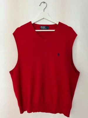 Röd väst från Polo Ralph Lauren - Klassisk röd väst från Polo Ralph Lauren med v-ringad hals och diskret blå logga broderad på bröstet. Västen har en enkel och stilren design som passar till många olika outfits. Tillverkad i mjukt bomullsmaterial för en bekväm känsla.