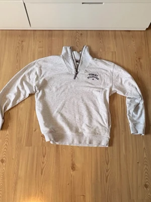 Ljusgrå half zip från Tommy Hilfiger - Ljusgrå långärmad half zip från Tommy Hilfiger med broderad logga på bröstet. Skön och avslappnad passform, perfekt till vardags. Tillverkad i mjuk bomullsmix och har ribbade muddar vid ärmslut och nederkant.