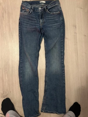 Blå raka jeans med fem fickor - Säljer ett par klassiska blå jeans med rak passform och fem fickor. Jeansen har normal midja och subtila slitningar för en avslappnad look. Tillverkade i denim med kontrastsömmar och knappgylf.