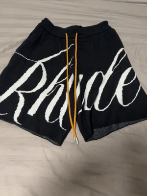Svarta shorts med vit text och snörning - Säljer ett par svarta shorts med stor vit text framtill och långa gula snören i midjan. Shortsen har elastisk midja och en bakficka. Perfekta för en avslappnad och trendig stil.