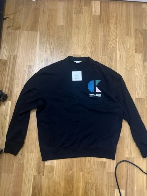 Svart sweatshirt från Calvin Klein - Svart sweatshirt från Calvin Klein med färgglad CK-logga i blått, grönt och rosa på bröstet. Tröjan har rund halsringning och ribbade muddar vid ärmslut och nederkant. Perfekt för en avslappnad och trendig stil.