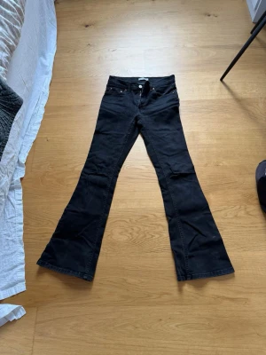 Svarta bootcut jeans med låg midja - Säljer ett par svarta bootcut jeans med låg midja och klassisk femficksmodell. Jeansen har utsvängda ben och silverfärgade detaljer vid fickorna. Materialet är stretchigt denim som sitter snyggt och bekvämt. Perfekta för dig som gillar retrovibbar och vill ha en cool siluett.