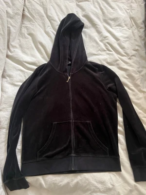 Svart velourhoodie med dragkedja - Mjuk och skön svart hoodie i velour med dragkedja framtill och känguruficka. Klassisk huva och ribbade muddar vid ärmslut och nederkant. Perfekt för en avslappnad och trendig look.