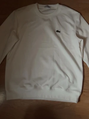  Lacoste - Klassisk vit sweatshirt från Lacoste med rund halsringning och den ikoniska krokodilloggan broderad på bröstet. Tröjan har långa ärmar och ribbade muddar vid ärmslut och nederkant. Perfekt för en clean och stilren look.