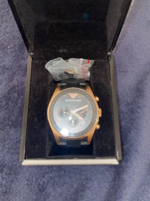 Emporio Armani svart/guld klocka - Stilren klocka från Emporio Armani med svart urtavla, guldfärgat boett och svarta länkar. Klockan har kronograf-funktion och datumvisning. Tillverkad i rostfritt stål och levereras i originalask. Perfekt accessoar för dig som gillar exklusiv design.