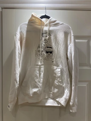 Vit hoodie med hiphop-tryck - Säljer en vit hoodie med huva och känguruficka. På framsidan finns ett svart tryck med tecknade porträtt av kända hiphop-artister och texten 'Compton'. Hoodien har dragsko i huvan och ribbade muddar vid ärmar och nederkant. Perfekt för dig som gillar streetwear.