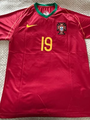 Portugal fotbollströja Nani 19 Nike - Säljer en röd Portugal fotbollströja från Nike med nummer 19 och Nani på ryggen. Tröjan har gula detaljer, grönt vid kragen och Portugals landslagsemblem på bröstet. Tillverkad i lätt och ventilerande material, perfekt för match eller träning.
