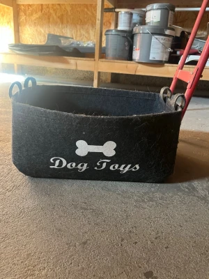 Svart filtbox för hundleksaker - Praktisk svart förvaringsbox i filt med texten 'Dog Toys' och ett benmotiv på framsidan. Perfekt för att samla hundens leksaker på ett ställe. Boxen har fyra öglor i hörnen för enkel hantering och är mjuk men stadig i formen.