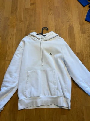 Vit hoodie med logga från Lacoste - Säljer en clean vit hoodie från Lacoste med klassisk krokodillogga på bröstet. Hoodien har stor ficka framtill, dragsko i huvan och ribbade muddar. Perfekt för dig som gillar enkel och stilren streetwear.