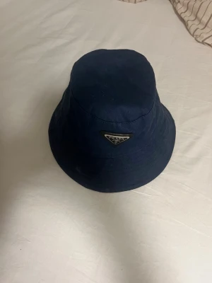 Mörkblå bucket hat från Prada - Snygg mörkblå bucket hat från Prada med klassisk triangellogo framtill. Hatten är tillverkad i ett slitstarkt tyg och har en avslappnad passform. Perfekt accessoar för att lyfta din stil och skydda mot solen.