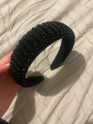Brett svart diadem med pärlor - Snyggt svart diadem med bred form, täckt av små svarta pärlor och glittrande stenar som ger en lyxig och trendig känsla. Perfekt accessoar för att lyfta din look och ge lite extra edge till håret. Diademet har en vadderad insida för bekväm användning.