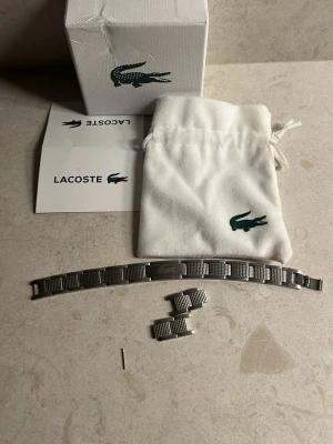 Lacoste silverfärgad klockarmband - Snyggt silverfärgat klockarmband från Lacoste i metall med klassisk länkdesign. Kommer med extra länkar, originalpåse och box. Perfekt för dig som vill byta ut eller uppgradera din klocka med en stilren och tidlös look.
