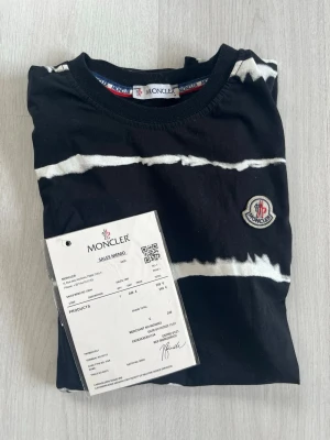 Svart och vit randig Moncler t-shirt - Snygg svart t-shirt från Moncler med vita oregelbundna ränder och klassisk Moncler-logga på bröstet. T-shirten har rund hals och är tillverkad i mjuk bomull som känns skön mot huden. Perfekt för dig som gillar streetwear och exklusiva märken.