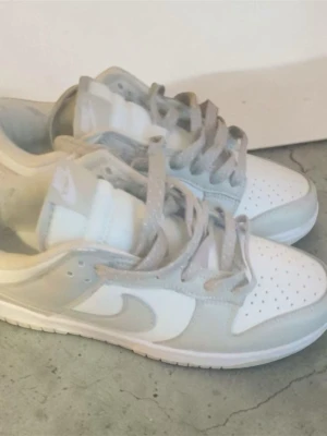 Nike Dunk Low vita och rosa sneakers - Nike Dunk Low sneakers i vitt och ljusrosa med klassisk swoosh på sidan. Ovandel i läder och textil, perforerad tå och platt sula. Broderad Nike-logga på hälen och matchande  glittrande rosa skosnören. Perfekta för en clean och stilren look.